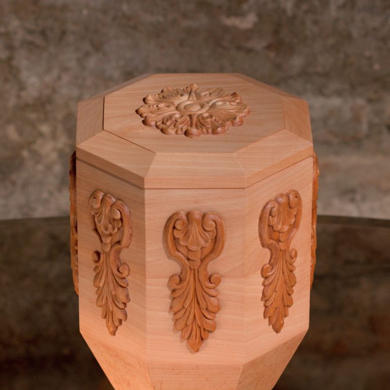 Holzurne mit sechseckigem Körper und Facettenschliff an den Längskanten, glatter Oberfläche. Die Urne verjüngt sich im unteren Bereich.