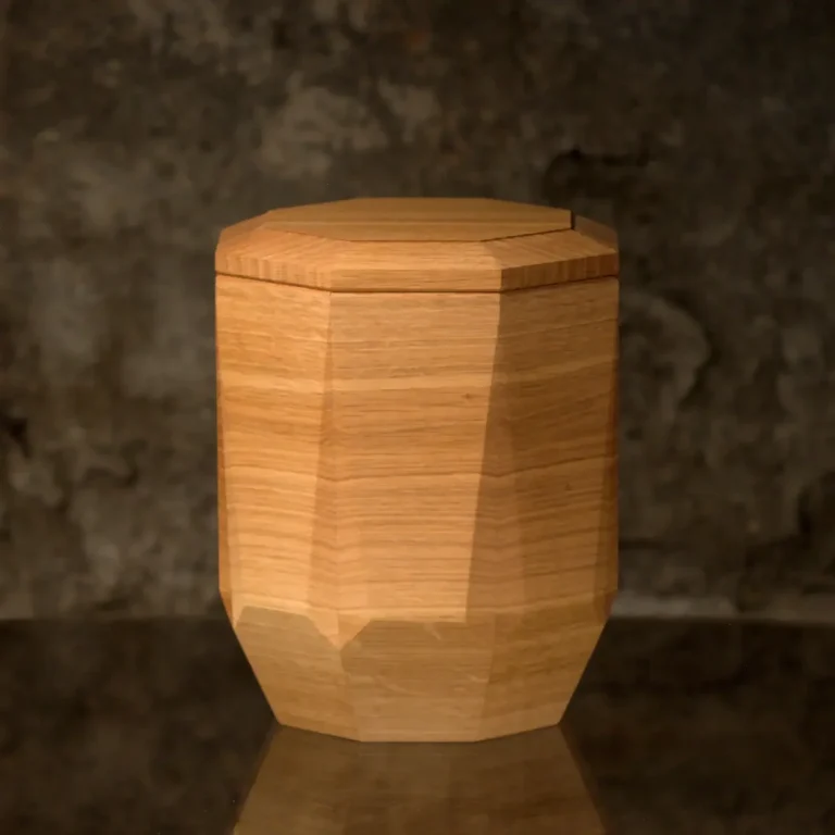 Holzurne mit sechseckigem Körper und Facettenschliff an den Längskanten, glatter Oberfläche. Die Urne verjüngt sich im unteren Bereich.