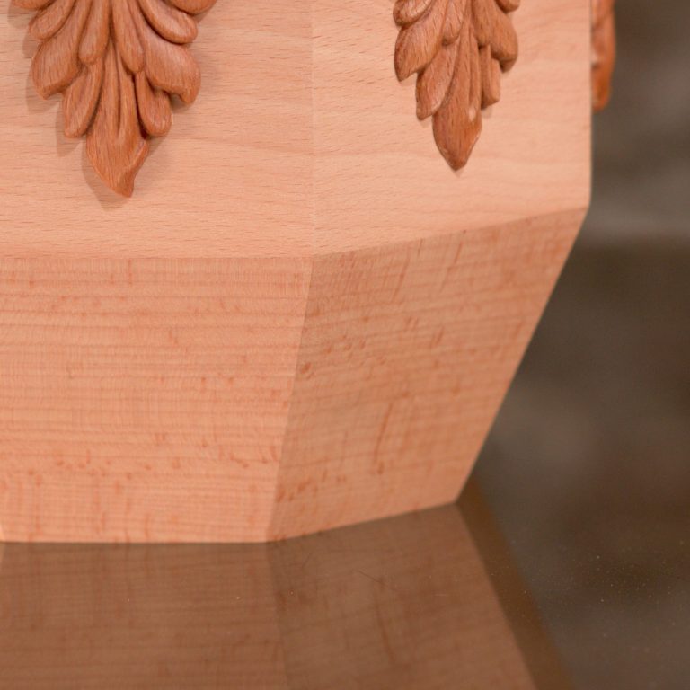 Holzurne mit sechseckigem Körper und Facettenschliff an den Längskanten, glatter Oberfläche. Die Urne verjüngt sich im unteren Bereich.