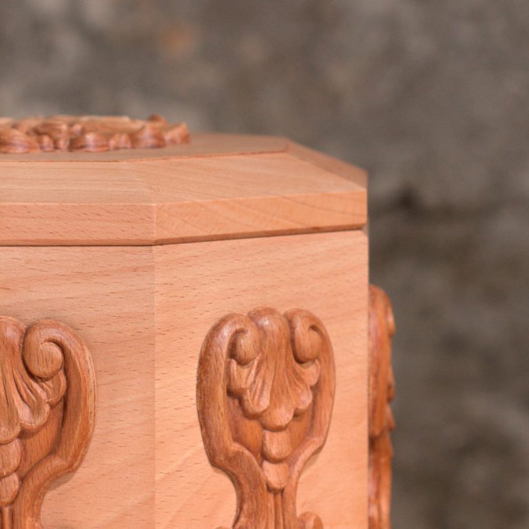 Holzurne mit sechseckigem Körper und Facettenschliff an den Längskanten, glatter Oberfläche. Die Urne verjüngt sich im unteren Bereich.